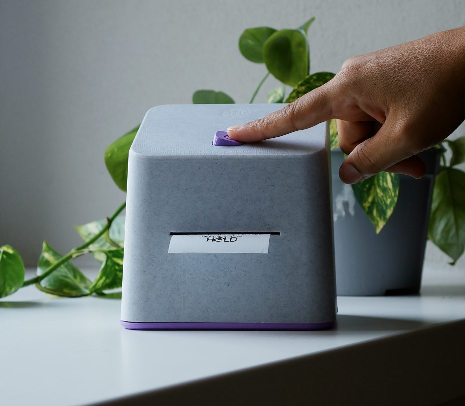 A hand pressing the button on top of the HELD device, next to a plant