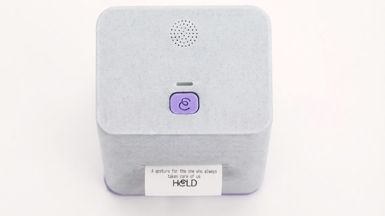 Top view of HELD with its button and a printed note reading 'A gesture for the one who always takes care of us'