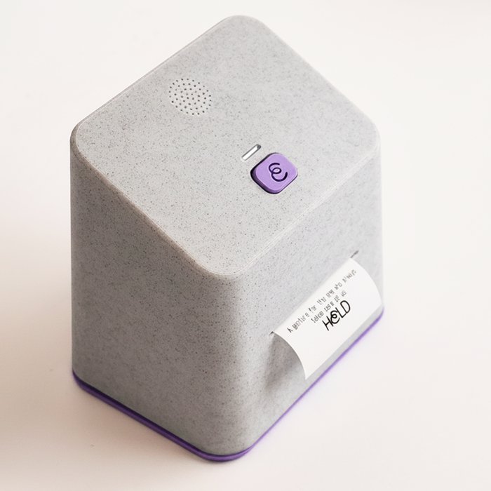 The HELD device, a soft grey cube with a button and a printed note